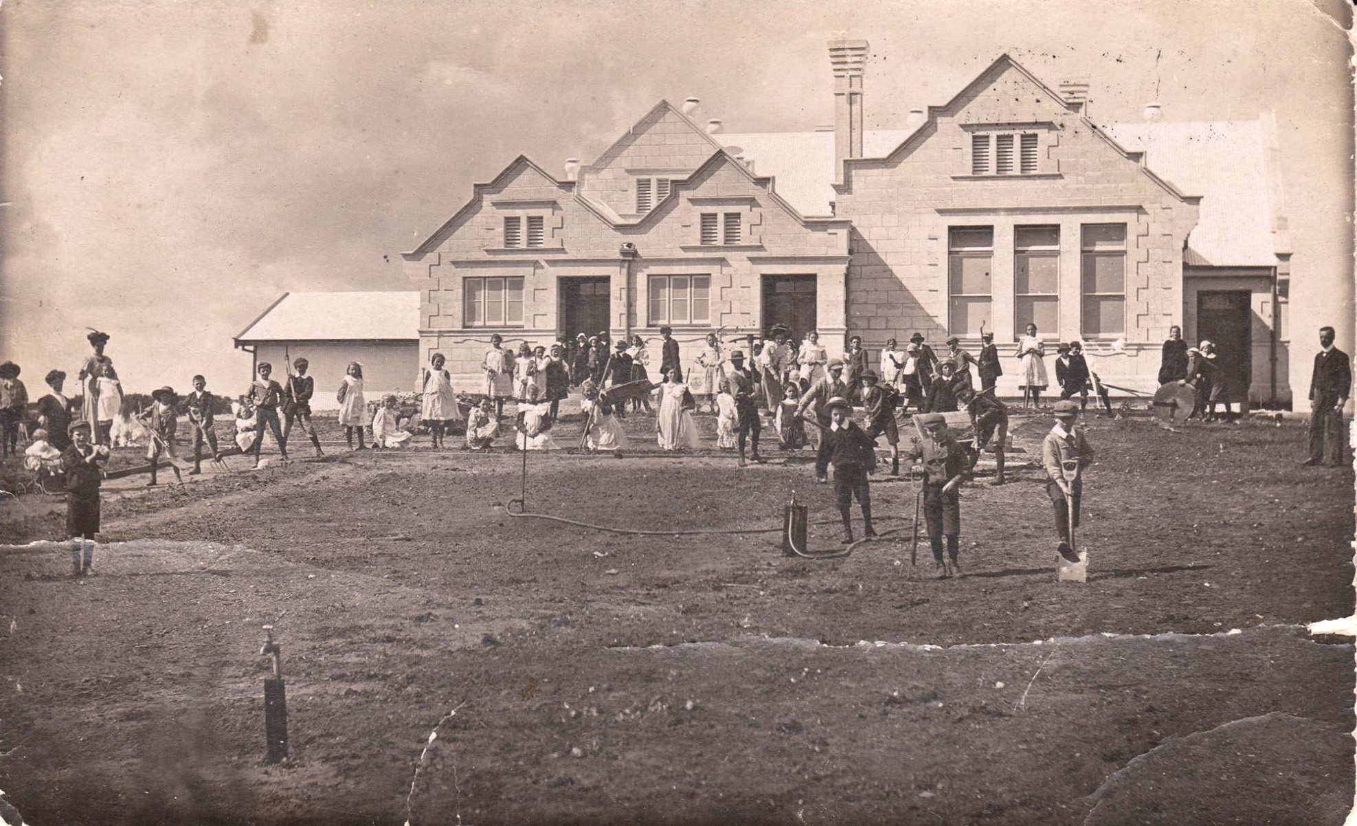 The image is a photo of a grand looking country primary school with four pitches to the roof and at least two chimneys. There are windows in groups of two’s and three’s. In front of the school are approximately 50 children dressed in olden day clothes and they are looking toward the camera. Some of the boys hold spades and rakes. The children are not in formation and have been working to establish the gardens. On either side of the photo are teachers or other adults.